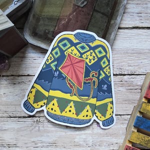 May include: A sticker featuring a blue sweater design with yellow and green accents. The sweater has a kite graphic in red and yellow. The sticker is on a wooden surface.