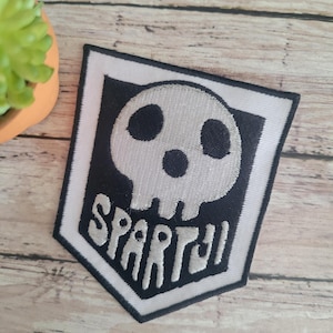 May include: White and black embroidered patch with a skull and the word "SPARTY" in a white font.