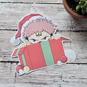 May include: A sticker featuring an anime character wearing a Santa hat, peeking over a red gift box with green stripes. The character has pink hair and red eyes. Decorative ornaments are scattered around the box.