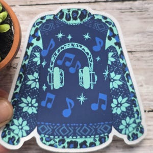 May include: A sticker featuring a navy blue sweater design with teal accents. The sweater has a pattern of snowflakes, musical notes, and a pair of headphones. The design is reminiscent of a festive holiday theme.