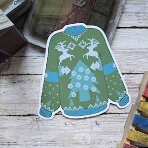 May include: A sticker of a green sweater with a blue collar and cuffs. The sweater features white reindeer and a Christmas tree design. Blue bands accent the sleeves. The sticker is on a wooden surface.