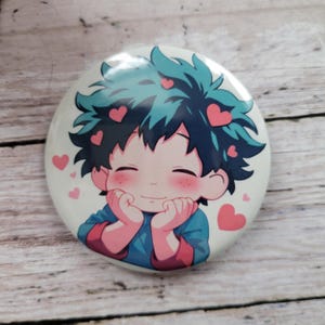 May include: A round button featuring a cartoon illustration of a character with green hair, blushing cheeks, and a big smile. The character is holding their hands to their face in a gesture of love. Pink hearts are scattered around the character's head.