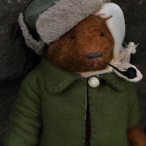 Può includere: Un orsacchiotto di peluche marrone che indossa una giacca verde, una sciarpa bianca e un cappello di pelliccia marrone e verde con le alette per le orecchie. L'orso tiene un tubo nero in bocca.