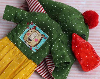 Puede incluir: Un vestido de muñeca verde y amarillo con lunares blancos y un ribete rojo y blanco. El vestido tiene un parche con la cara de un oso de dibujos animados en la parte delantera. La muñeca también tiene un sombrero rojo con un pompón blanco.