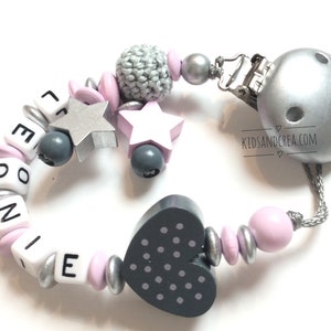 Puede incluir: Un mordedor para bebé rosa y gris con el nombre Leonie escrito en letras blancas. El mordedor tiene una cuenta de madera en forma de corazón con lunares blancos, un clip plateado y una cuenta de crochet gris.