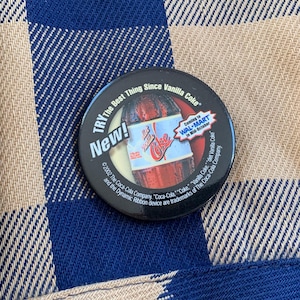 May include: A black and white button with the text "TRY The Best Thing Since Vanilla Coke" and "New!" in a red circle. The button also features a Coca-Cola bottle with the text "Coming to WAL-MART in Mid-October".