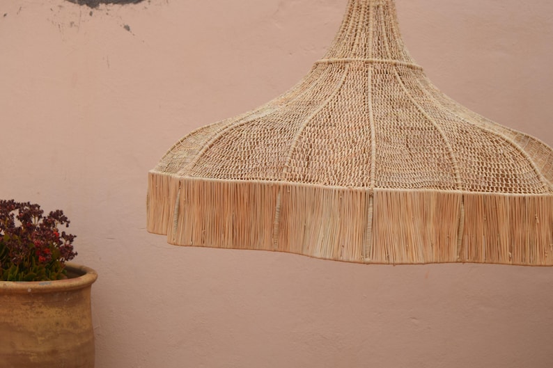 Op de afbeelding: Geweven hanglamp in natuurlijk beige met een gefranjerde rand, hangend voor een lichtroze muur. Linksonder is een terracotta pot met planten te zien. De lamp is een decoratief element.