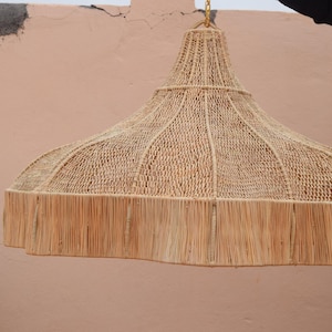 Op de afbeelding: Een grote, beige geweven lampenkap met een gefranjerde onderkant. De lampenkap heeft een klokvorm met een geweven bovenkant en een bredere, gefranjerde onderkant. Hij hangt aan een gouden ketting tegen een perzikkleurige muur.