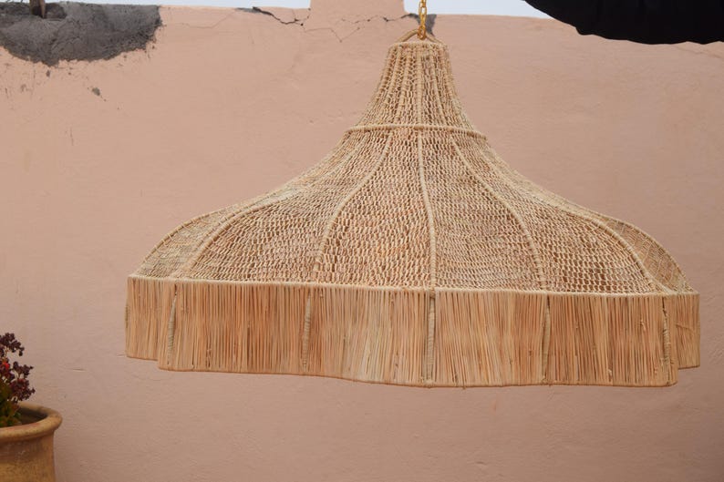 Op de afbeelding: Een grote, beige geweven lampenkap met een gefranjerde onderkant. De lampenkap heeft een klokvorm met een geweven bovenkant en een bredere, gefranjerde onderkant. Hij hangt aan een gouden ketting tegen een perzikkleurige muur.