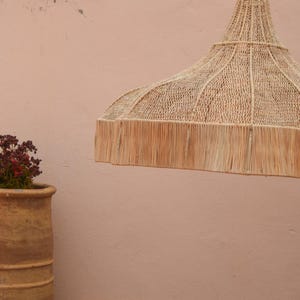 May include: A woven, natural fibre pendant light with a fringed edge hangs against a peach-coloured wall. A large, aged terracotta pot with a plant sits to the left. A window with a metal frame is visible in the background.