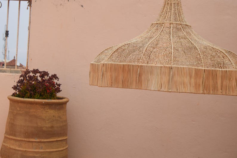 May include: A woven, natural fibre pendant light with a fringed edge hangs against a peach-coloured wall. A large, aged terracotta pot with a plant sits to the left. A window with a metal frame is visible in the background.
