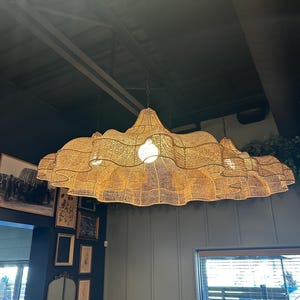 Può includere: Un grande lampadario in rattan intrecciato è appeso al soffitto. L'apparecchio ha una forma ondulata unica ed emette un bagliore caldo e invitante. L'apparecchio è sospeso da catene ed è un pezzo forte.