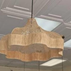 Op de afbeelding: Een geweven, beige hanglamp met een gelaagd, geschulpt ontwerp. Het licht hangt aan een zwart snoer en is opgehangen aan een wit plafond. De armatuur heeft een natuurlijk, getextureerd uiterlijk.
