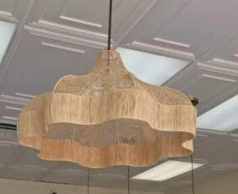 Op de afbeelding: Een geweven, beige hanglamp met een gelaagd, geschulpt ontwerp. Het licht hangt aan een zwart snoer en is opgehangen aan een wit plafond. De armatuur heeft een natuurlijk, getextureerd uiterlijk.