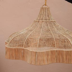 Peut inclure: Un luminaire suspendu tissé avec un bord festonné. Le luminaire est fait de fibres naturelles de couleur beige et est suspendu à une chaîne dorée. Le luminaire est suspendu devant un mur rose clair.