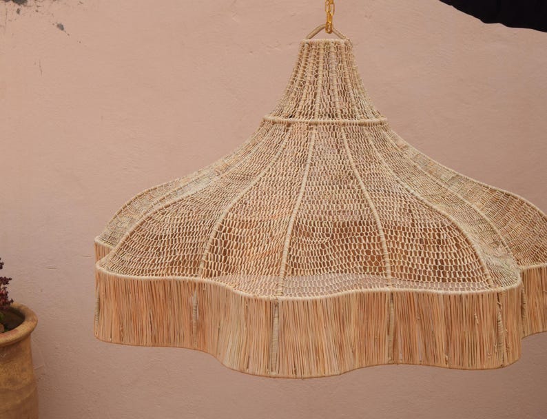 Op de afbeelding: Een geweven hanglamp met een geschulpte rand. De lamp is gemaakt van natuurlijke vezels in tinten beige en bruin. Het licht hangt aan een goudkleurige ketting en tegen een lichtroze muur.