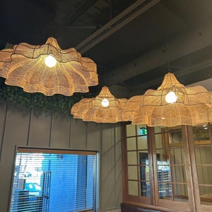 Op de afbeelding: Drie geweven hanglampen met een geschulpte rand hangen aan het plafond. De lampen hebben een warme, natuurlijke kleur en een gloeilamp in het midden. De lampen hangen in een restaurant of café.