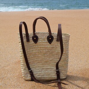 May include: A woven straw tote bag with brown leather straps and handles. The bag has a flap closure with a leather buckle and is sitting on a sandy beach.