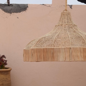 May include: A woven pendant light made from natural fibres, with a fringed edge, hangs against a peach-coloured wall. A terracotta pot with a plant sits to the left of the light. The light is suspended from a gold chain.