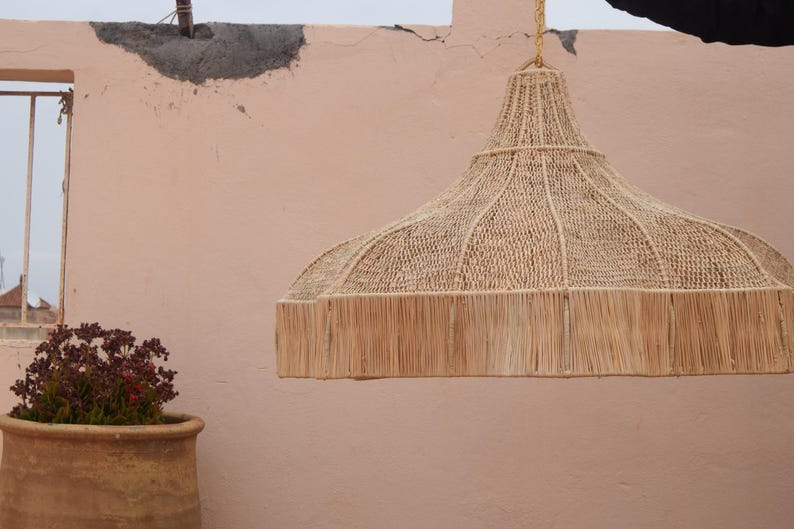 May include: A woven pendant light made from natural fibres, with a fringed edge, hangs against a peach-coloured wall. A terracotta pot with a plant sits to the left of the light. The light is suspended from a gold chain.