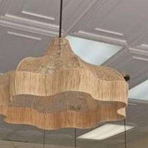 Op de afbeelding: Een beige geweven hanglamp met een geschulpte rand en een gefranjerde onderkant, opgehangen aan een zwart snoer. De lamp hangt aan een wit plafond.