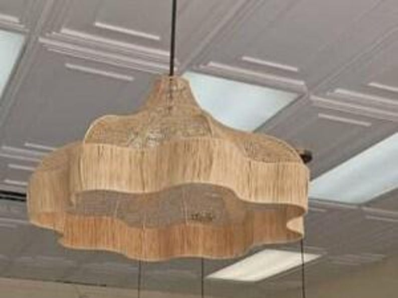 Op de afbeelding: Een beige geweven hanglamp met een geschulpte rand en een gefranjerde onderkant, opgehangen aan een zwart snoer. De lamp hangt aan een wit plafond.