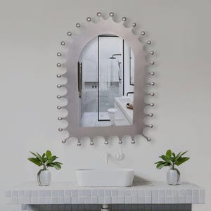 Może przedstawiać: Dekoracyjne, łukowate lustro łazienkowe z falistą krawędzią i małymi, srebrnymi kulkami. Lustro jest zamontowane nad białym prostokątnym zlewem i półką z kafelkami. Dwie małe rośliny doniczkowe flankują zlew.