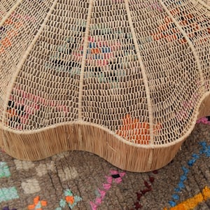 May include: A light brown, woven coffee table with a scalloped edge. The table features a woven top and a solid, fringed edge. It rests on a patterned rug with pink, orange, and blue accents.
