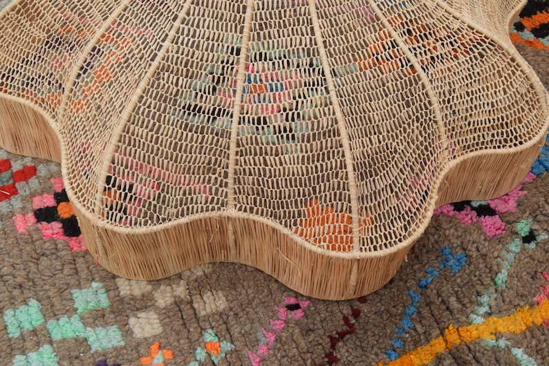 May include: A light brown, woven coffee table with a scalloped edge. The table features a woven top and a solid, fringed edge. It rests on a patterned rug with pink, orange, and blue accents.