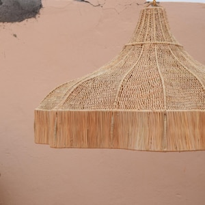 May include: A woven pendant light in natural beige. The bell-shaped shade has a fringed edge. The light hangs from a gold chain. The background is a light peach-coloured wall. The light would suit a living room or dining room.