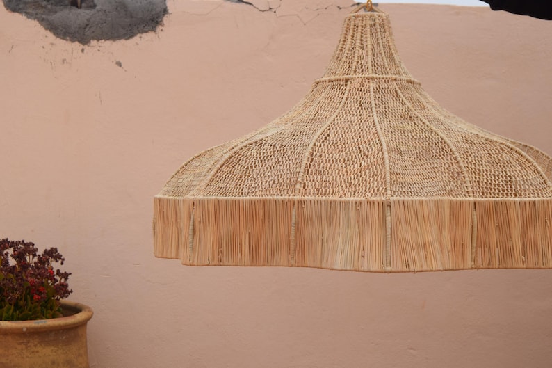 May include: A woven pendant light in natural beige. The bell-shaped shade has a fringed edge. The light hangs from a gold chain. The background is a light peach-coloured wall. The light would suit a living room or dining room.