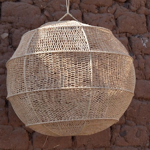 May include: A large, spherical woven basket with a light tan color, hanging against a textured, reddish-brown brick wall. The basket has a lattice-like design, and is suspended by a rope. The overall aesthetic is rustic and natural.