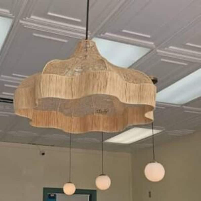 Op de afbeelding: Een geweven, beige hanglamp met een geschulpte rand en drie hangende gloeilampen. De lamp hangt aan een donker snoer en is bevestigd aan een wit plafond met een patroon. De gloeilampen zijn gelijkmatig verdeeld.