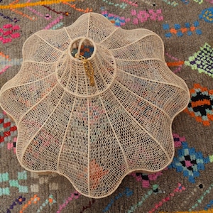 Op de afbeelding: Een cr&egrave;mekleurige, geweven hanglamp met een geschulpte rand, opgehangen aan een gouden ketting. Het licht staat op een grijze vloerkleed met een kleurrijk geometrisch patroon, met roze, oranje en blauwe accenten.