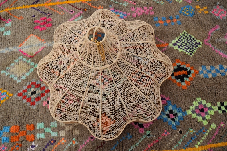 Op de afbeelding: Een cr&egrave;mekleurige, geweven hanglamp met een geschulpte rand, opgehangen aan een gouden ketting. Het licht staat op een grijze vloerkleed met een kleurrijk geometrisch patroon, met roze, oranje en blauwe accenten.