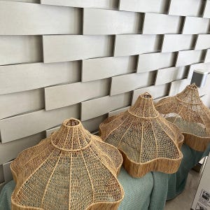 Peut inclure: Trois abat-jours en rotin tressé avec un design unique en forme d'éclat d'étoile. Les abat-jours sont de couleur beige naturel et ont un bord effiloché. Ils sont présentés contre un mur texturé gris clair.