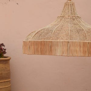 May include: A natural-toned woven pendant light, featuring a wide, rounded shade with a fringed edge. A terracotta pot with a plant is in the foreground. A window is visible in the background, set against a pale pink wall.