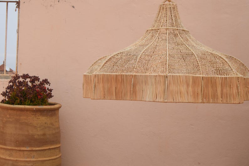 May include: A natural-toned woven pendant light, featuring a wide, rounded shade with a fringed edge. A terracotta pot with a plant is in the foreground. A window is visible in the background, set against a pale pink wall.