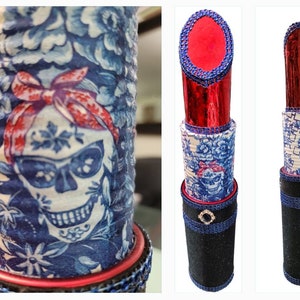 May include: Decorative lipstick-shaped container with a Day of the Dead skull design. The container features a blue and white floral pattern with a red bow and skull. The lipstick top is red and the base is black with a decorative jewel.