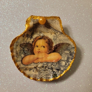May include: A gold-rimmed seashell ornament featuring a cherub image and a pearl accent. The cherub has wings and is holding its hands together. The seashell has a vintage-inspired design with musical notes and floral patterns.