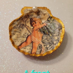 May include: A seashell ornament with a gold trim and a pearl on top. The seashell is decorated with a vintage image of a cherub holding a bouquet of flowers. The image is surrounded by a decorative border and text.