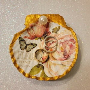 May include: A decorative seashell ring dish with a gold-painted edge. The dish features a floral design with a butterfly and the text "FLEUR PONT". Two rings and a pearl are on the dish.