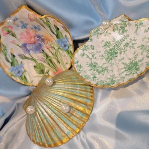 Peut inclure: Trois coquillages décoratifs. L'un présente un motif floral avec des fleurs roses, violettes et bleues. Un autre a un motif vert et blanc, et le troisième est doré et turquoise avec des fausses perles.