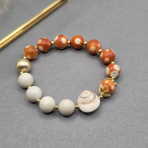 May include: A stretch bracelet featuring a series of round beads in orange with white spots and solid white beads. The design includes a single textured gold-tone bead, small gold-tone spacer beads, and a central white spiral seashell charm.