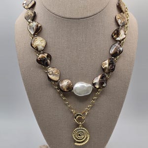 May include: A statement necklace featuring large, irregularly shaped brown and cream-colored beads connected by a gold chain. A large, white pearl is centered. A gold pendant in the shape of a spiral seashell hangs below.