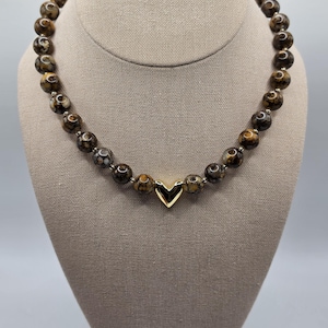 May include: A beaded necklace featuring round, mottled brown and tan stones with a polished finish. The beads are strung together with small metallic spacers, and a single, smooth, gold-toned heart-shaped charm is positioned at the center.