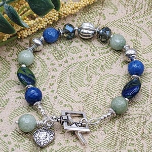 May include: A beaded bracelet featuring a mix of blue, green, and silver beads. The bracelet includes round and oval beads, with a heart charm and a toggle clasp. The beads are strung on a flexible cord.