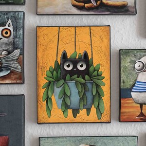 Puede incluir: Un gato negro con ojos grandes está colgado en una maceta azul llena de plantas verdes. El gato está suspendido del techo por dos cuerdas. El fondo es un lienzo amarillo con un diseño de mapa vintage.
