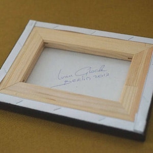 May include: A wooden frame with a white background and a handwritten signature that reads "Ivan Glock Berlin 2012".