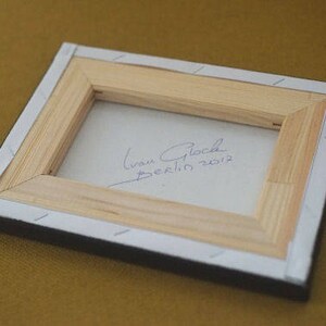 May include: A wooden frame with a white background and a handwritten signature that reads "Ivan Glock Berlin 2012".